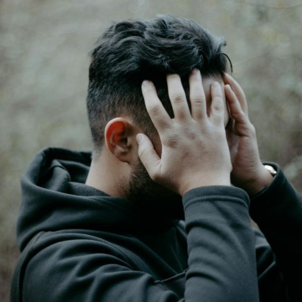 A person in a dark hoodie stands outdoors, hands covering their face, conveying distress. The background is blurred, creating an introspective mood.