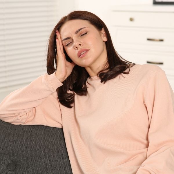 A woman in a peach sweater sits on a gray couch, holding her head in apparent discomfort. Her eyes are closed, conveying a sense of pain or fatigue.