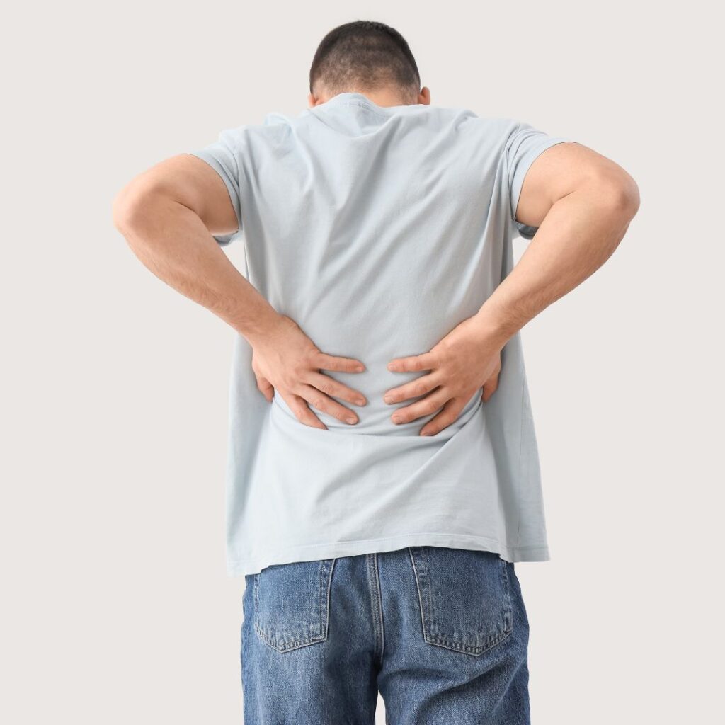 A man in a light blue t-shirt and jeans stands with his back to the camera, holding his lower back with both hands, suggesting discomfort or pain.