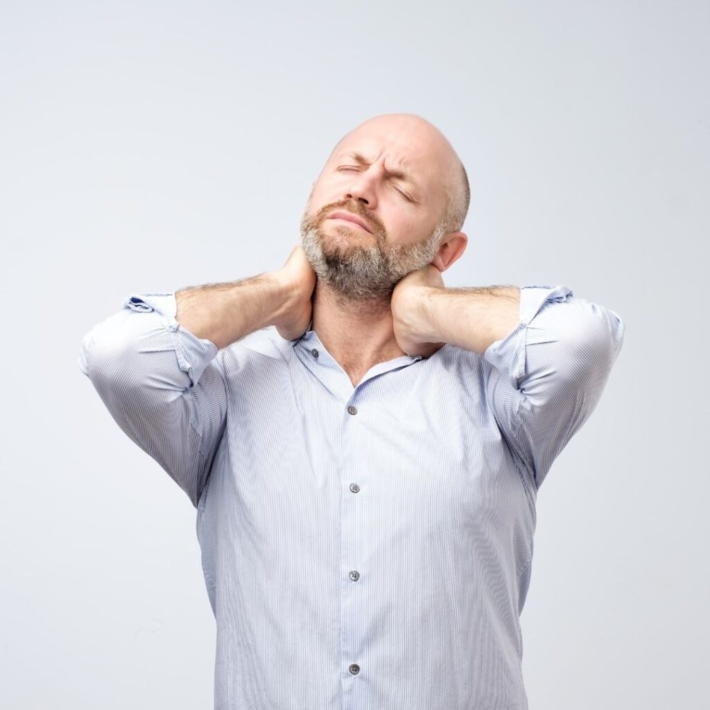 A bearded man in a striped shirt massages his neck, eyes closed in discomfort, against a plain background, conveying tension and relief.
