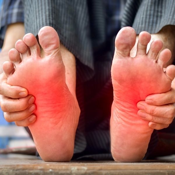 Close-up of a person holding their feet, showing redness on the soles, indicating discomfort or pain. The background shows a checkered fabric.