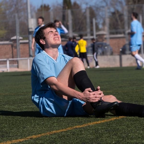 A soccer player in a blue uniform sits on a field, grimacing in pain while holding his ankle. Teammates in the background continue playing.