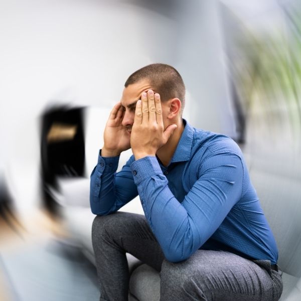 A man in a blue shirt sits with his elbows on his knees, pressing his fingers to his temples. The background is blurred, conveying stress or dizziness.